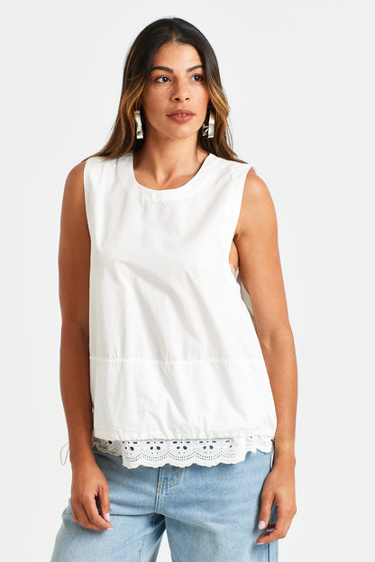 AUGUSTE TOP Woman wearing a white sleeveless top with lace trim and light blue jeans on a white background
